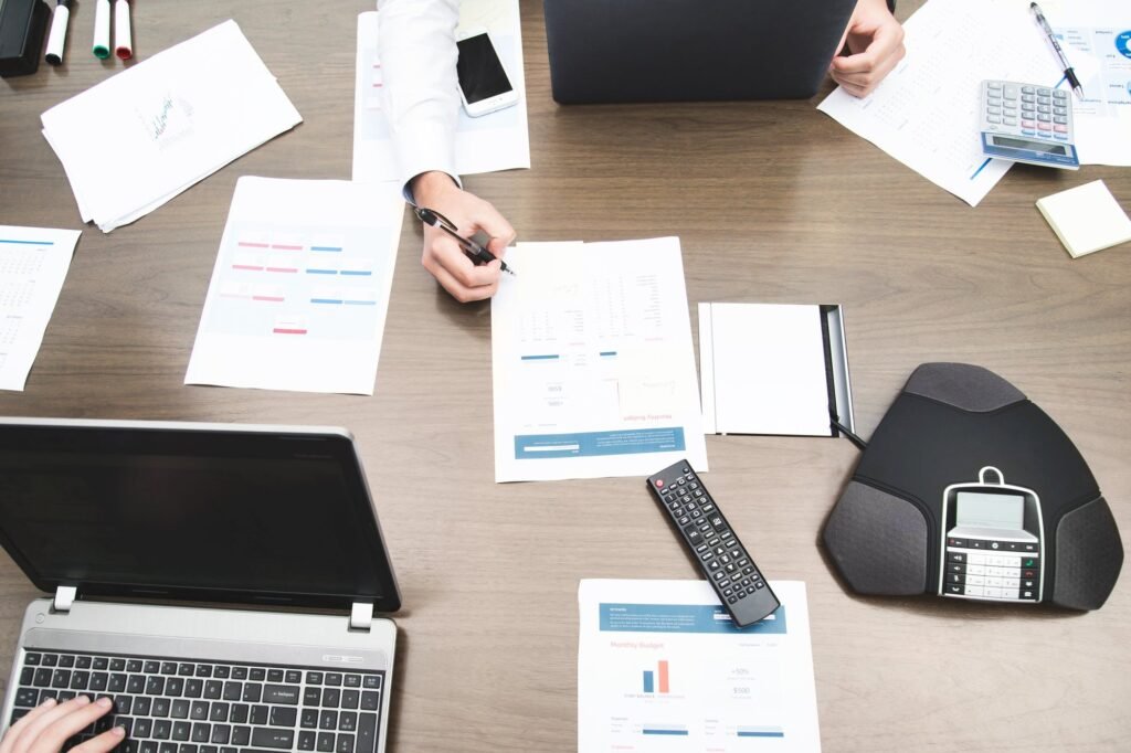 Overhead view of a meeting table with printed charts and reports, two laptops, a notebook and a conference speakerphone.