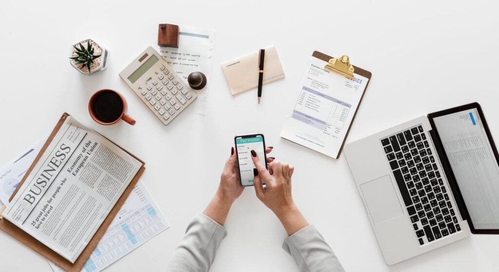 Overhead view of a white desk with a calculator, a clipboard holding an invoice, a notebook, a laptop, a cup of coffee and a phone held in two hands.